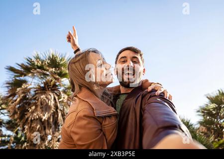 Glückliches Paar, das draußen ein fröhliches Selfie mit Palmen und blauem Himmel macht. Sie lächeln und umarmen sich, halten einen Moment unbeschwerten Spaß und Ex fest Stockfoto