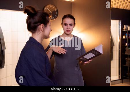 Eine junge Frau im Bademantel lächelt und wählt eine Creme im Spa. Die Kosmetikerin bietet ihrem attraktiven Kunden verschiedene Gesichtspflegeprodukte an Stockfoto