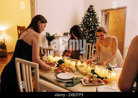 Junge attraktive Frauen in Abendkleidern stellen den festlichen Tisch. Charmante Mädchen zünden Kerzen für eine Party an. Ein geschmückter Weihnachtsbaum und ein gemütliches Leben Stockfoto