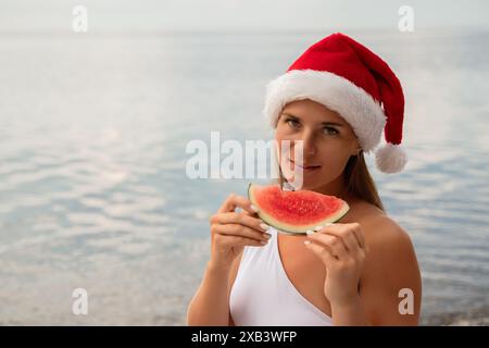 Eine Frau isst eine Wassermelone, während sie einen roten weihnachtsmann-Hut trägt. Die Szene ist unbeschwert und festlich, während die Frau einen erfrischenden Snack auf einem sunn genießt Stockfoto