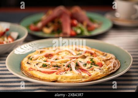 Köstliches großes Omelett mit Speck und Tomaten wird zum Frühstück auf dem Tisch serviert. Hausmannskonzept Stockfoto