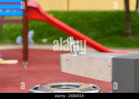 Moderner Chrom-Wasserbrunnen auf einem Spielplatz mit einer rot-blauen Rutsche im Hintergrund. Konzept der öffentlichen Barrierefreiheit Stockfoto