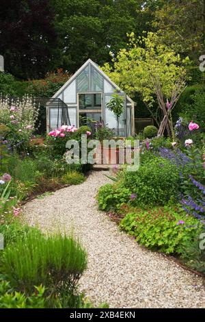 Gewächshaus in einem englischen Landgarten - Schotterweg schlängelt sich durch lebendige Blumen und Sträucher, die zu einem bezaubernden Gewächshaus führen. Stockfoto