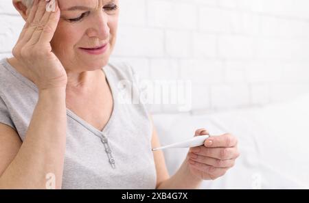 Frau, die das Thermometer in der Hand hält und den Kopf berührt Stockfoto