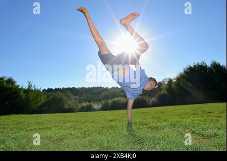 Ein Junge, der einen Handstand auf einem grasbewachsenen Feld macht, während die Sonne hell am klaren blauen Himmel in Bayern scheint Stockfoto