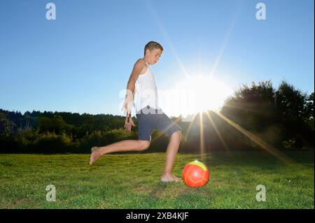 Ein Junge, der einen Fußball auf einem grasbewachsenen Feld tritt, während die Sonne im Hintergrund untergeht, Bayern Stockfoto