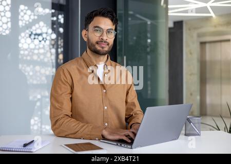 Ein selbstbewusster junger Mann mit Brille und braunem Hemd arbeitet an seinem Laptop an einem Schreibtisch in einem stilvollen, modernen Büro. Stockfoto