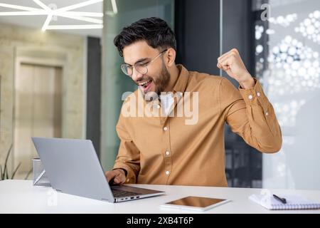 Glücklicher junger muslimischer Mann, der im Büro am Tisch sitzt, auf den Laptop-Bildschirm blickt und Erfolge und Erfolge genießt, mit der Hand Siegesgesgesen macht ja. Stockfoto