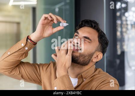 Nahaufnahme eines jungen Indianers, der in einem modernen Büro steht und ihm Medikamente gegen Erschöpfung und Entzündung ins Auge gibt. Stockfoto