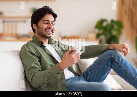 Ein glücklicher, gutaussehender Mann, der das Smartphone benutzt, während er auf Einer Couch sitzt Stockfoto