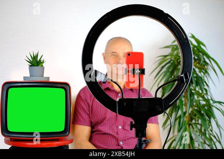 Charmanter Mann mittleren Alters, 60-65 Jahre alter Blogger sitzt vor Ringlicht und rotem Smartphone und spricht emotional über das Thema Wirtschaft, Politik Dur Stockfoto