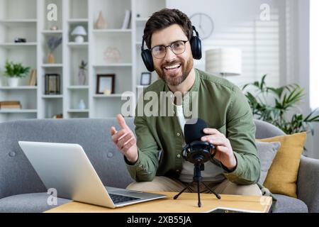 Porträt eines lächelnden jungen Mannes, der von zu Hause aus arbeitet, Kopfhörer trägt, auf einem Sofa vor einem Laptop und einem Mikrofon sitzt und mit den Händen zur Kamera spricht und Gesten macht. Stockfoto