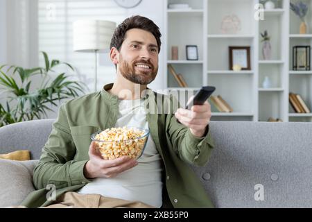 Ein lächelnder und entspannter junger Mann sitzt zu Hause auf der Couch und entspannt sich vor dem Fernseher, isst Popcorn und schaltet mit der Fernbedienung um. Stockfoto