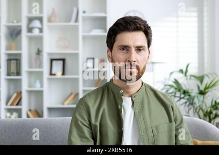 Nahporträt eines ernsten jungen Mannes in einem grünen Hemd, der zu Hause auf der Couch sitzt und in die Kamera schaut. Stockfoto