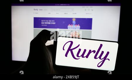 Person, die ein Mobiltelefon mit dem Logo des südkoreanischen E-Commerce-Unternehmens Kurly vor der Business-Webseite hält. Schwerpunkt auf dem Display des Telefons. Stockfoto