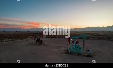 Pumpjack und Sendetürme bei Sonnenuntergang symbolisieren die Energiewende. Ein Pumpenheber pumpt Öl aus einem Brunnen mit Silhouetten von Strommasten Stockfoto