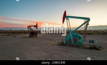 Pumpjack und Sendetürme bei Sonnenuntergang symbolisieren die Energiewende. Ein Pumpenheber pumpt Öl aus einem Brunnen mit Silhouetten von Strommasten Stockfoto