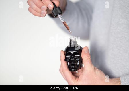 Junger Mann, der Bartöl-Glasflasche mit Pipette in der Nähe hält, schöner Mann, der kosmetisches Serum mit Pipette aufträgt, schwarzer Schädel. Essential Oder Cannabis Stockfoto