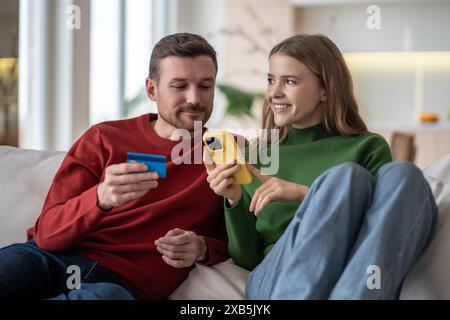 Online-Shopping zu Hause. Interessierte Paare bezahlen für Einkäufe mit Bankkarte, das Telefon sitzt auf der Couch. Stockfoto
