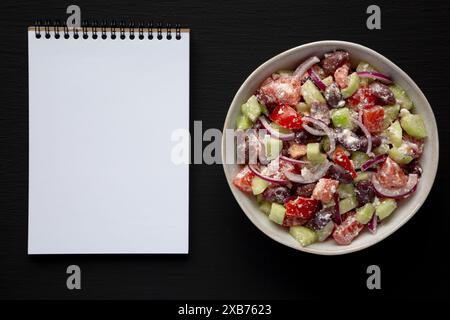 Hausgemachter mediterraner Salat in einer Schüssel, leerer Notizblock auf schwarzem Hintergrund, Draufsicht. Stockfoto