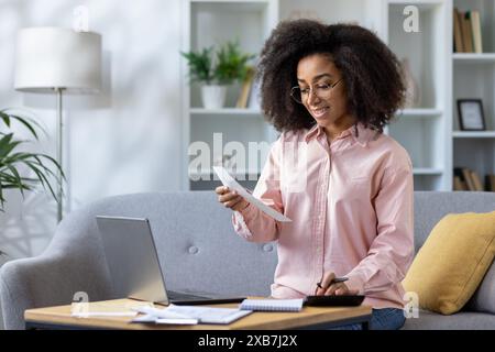 Eine junge Frau mit lockigen Haaren lächelt, während sie von zu Hause aus arbeitet, Dokumente liest und einen Laptop auf einer Couch benutzt. Stockfoto