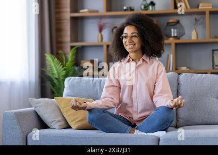 Lächelnde Frau übt Yoga und Meditation auf einem gemütlichen Sofa zu Hause. Entspannte, komfortable und zufriedene Atmosphäre in einem ruhigen Innenbereich. Stockfoto