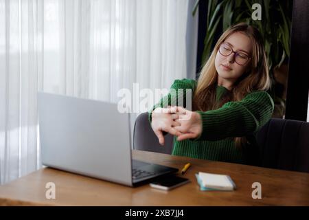 Eine Frau sitzt an einem Schreibtisch vor einem Laptop und streckt ihre Arme. Stichwörter Tisch, Computer, Laptop, Holz, Geste, PC, F Stockfoto