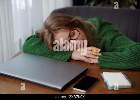 Eine Frau in einem grünen Pullover schläft friedlich auf einem Schreibtisch neben einem Laptop in einem Büro Stockfoto