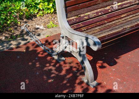 Holzbank im Park, mit einer Metallkette an einem Bolzen in den Boden gekettet. Stockfoto