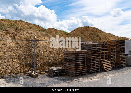 Holzpaletten stapeln sich vor einem Metallzaun vor dem Hintergrund großer Erdhaufen unter einem leicht bewölkten Himmel. Stockfoto