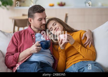 Ein zärtlicher Moment, ein familienliebendes Paar mit Umarmung. Entspannter Mann und Frau umarmen sich auf der Couch mit Tassen Kaffee Stockfoto