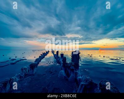 Ein ruhiger Sonnenuntergang über einem ruhigen Meer mit einem alten, verwitterten Holzpier, der sich unter dramatischem Himmel ins Wasser erstreckt. Stockfoto