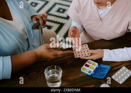 Junge Krankenschwester demonstriert Pillen bei afroamerikanischer Patientin, die mit Pillen-Kisten und einem Glas Wasser am Holztisch sitzt Stockfoto