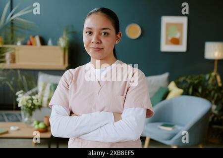 Die junge Krankenschwester in Rosenschnecken lächelt leicht und kreuzt ihre Arme, während sie für ein Porträt posiert Stockfoto