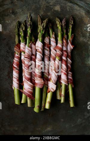 Mit Speck umwickelter Spargel in einem rustikalen, dunklen Tablett angeordnet. Der Spargel ist frisch und grün, eng mit herzhaftem Speck umwickelt, was eine optisch anmutende Tüte ergibt Stockfoto