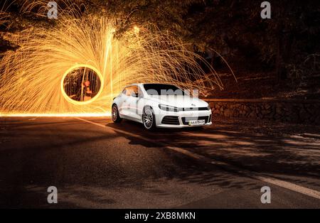 Spinnende Stahlwolle hinter weißem Volkswagen Scirocco. Dreiviertelansicht des VW-Fließhecks bei Nacht und Funken, die hinten in alle Richtungen herausfliegen. Stockfoto