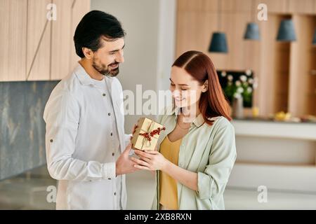 Eine rothaarige Frau und ein bärtiger Mann teilen sich eine Geschenkbox in einer modernen Küche, tauschen Lächeln aus und verbreiten Freude. Stockfoto