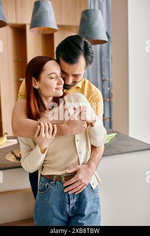 Eine rothaarige Frau und ein bärtiger Mann umarmen sich zärtlich in ihrer modernen Küche und teilen einen Moment der Liebe und Verbindung. Stockfoto