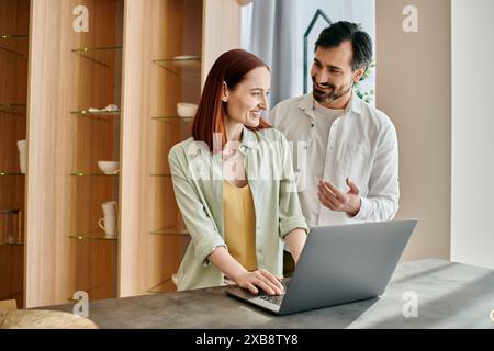 Eine rothaarige Frau und ein bärtiger Mann, der in einer stilvollen Küche mit einem Laptop beschäftigt ist, tief in Gesprächen und Verbundenheit. Stockfoto