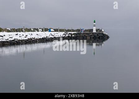 Ein einsamer Leuchtturm steht auf einem schneebedeckten Wellenbrecher, dessen grünes Licht an einem nebeligen Wintertag im ruhigen Wasser des Hafens reflektiert. Stockfoto