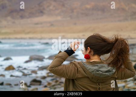 Eine junge Frau mit einer roten Blume im Haar steht an einer felsigen Küste und passt ihre Sonnenbrille an Stockfoto