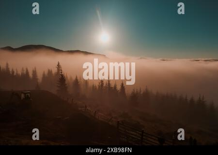 Ein nebeliger Morgen in den Bergen, bei dem die Sonne durch die Wolken schaut und ein warmes Leuchten über die Landschaft wirft. Die Silhouette von Waldbäumen und Hügeln ist in der Ferne sichtbar. Karpaten Stockfoto