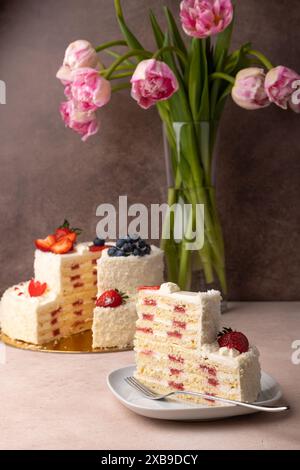 Erdbeeren-Kokos-Geburtstagskuchen mit Sahne und frischen Beeren. Ein Stück hausgemachter, mehrschichtiger Geburtstagskuchen. Im Hintergrund ist ein Strauß aus rosa t Stockfoto