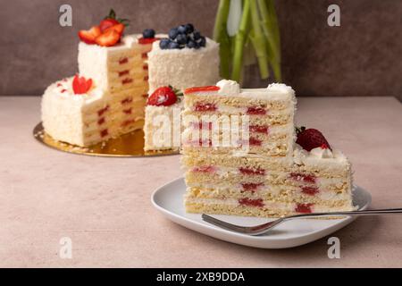 Erdbeeren-Kokos-Geburtstagskuchen mit Sahne und frischen Beeren. Ein Stück hausgemachter, mehrschichtiger Geburtstagskuchen. Im Hintergrund ist ein Blumenstrauß. Stockfoto