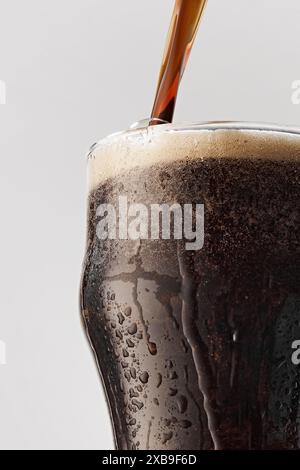 Das warme, dunkle Craft-Bier wird in einen Becher mit Kondenswasser auf Glas vor grauem Studiohintergrund gegossen. Stockfoto