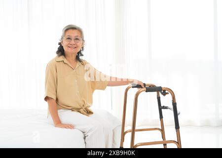 Sie lächelt und ist glücklich. Eine Frau in einem braunen Hemd sitzt auf einem Bett mit einem Walker neben ihr. Stockfoto