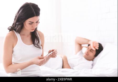 Frau Überprüft Thermometer, Während Der Mann Im Bett Liegt Stockfoto