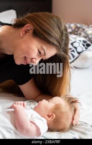 Die fürsorgliche junge Mutter spielt mit ihrer neugeborenen Tochter im Bett und sieht sie mit einem Lächeln an, glückliche Momente der Schwangerschaft Stockfoto