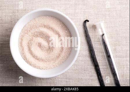 Vanillezucker, aromatisierter Zucker in einer weißen Schüssel auf Leinengewebe, neben getrockneten, braunen, Reifen Schoten oder Bohnen von Vanilla planifolia, einem Gewürz. Stockfoto