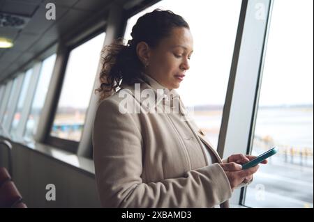 Geschäftsfrau, die am Fenster des Flughafenterminals steht und auf ihrem Smartphone tippt. Sie scheint konzentriert zu sein und liest möglicherweise E-Mails oder Nachrichten, während sie wartet Stockfoto